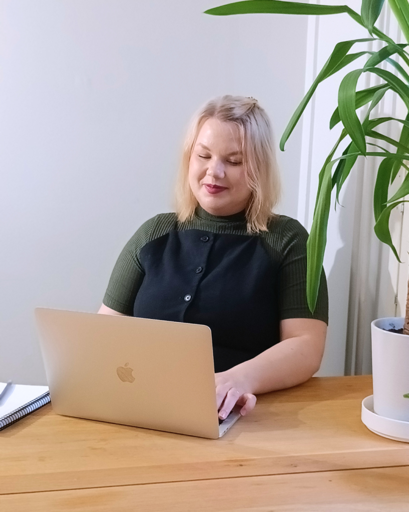 Nainen istuu pöydän takana ja hymyillen näpyttelee kannettavaa tietokonetta.