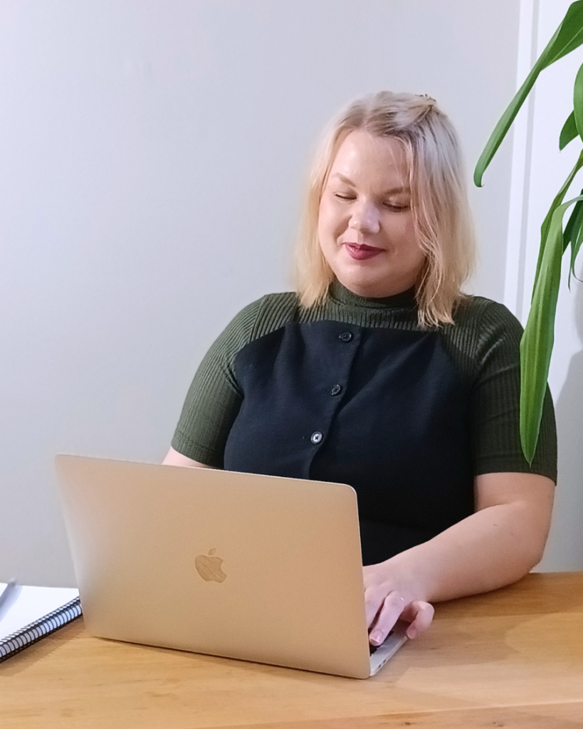 Nainen istuu hymyillen pöydän ääressä ja käyttää kannettavaa tietokonetta.
