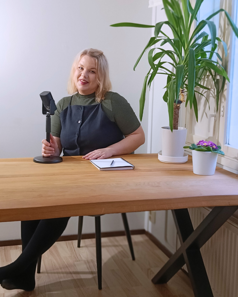 Nainen istuu puisen pöydän takana, jonka päällä on mikrofoni, vihko ja kasveja. Nainen selittää jotain pidellen mikrofonista kiinni.